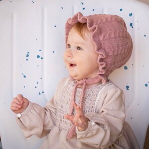 May include: A baby girl wearing a pink knitted bonnet with a ruffled edge and a cream-colored dress with a smocked bodice and gold accents.