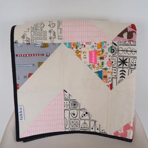 Może przedstawiać: Kołdra patchworkowa z różnorodnymi wzorzystymi tkaninami. Kołdra posiada geometryczne kształty i mieszankę kolorów, w tym różowy, szary i kremowy. Kołdra ma czarną obwódkę i etykietę z napisem "lukku".