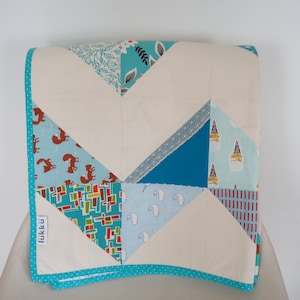 Puede incluir: Una colcha de bebé blanca con un patrón de chevron hecho de diferentes retazos de tela. La colcha tiene un borde turquesa con lunares blancos y una pequeña etiqueta que dice "lukkü".