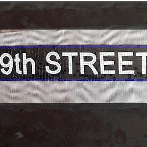 May include: A mosaic tile sign that reads "49th STREET" in white letters against a black background with a blue border. The sign is made of small, square tiles and is likely for a street or subway station.