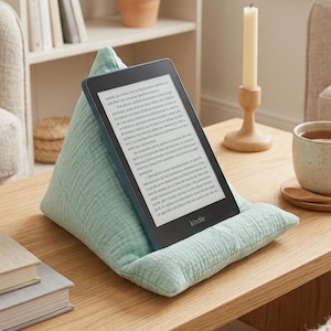Peut inclure: Un lecteur Kindle est posé sur un coussin vert clair sur une table basse en bois. Une tasse de thé, une bougie et des livres sont également sur la table. L'écran du Kindle affiche du texte. Le mot "Kindle" est visible sur l'appareil.