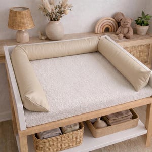 Op de afbeelding: Een witte houten commode met een crèmekleurige aankleedkussen en een crèmekleurige bumper. De tafel heeft twee rieten manden op de onderste plank. Een lamp, vaas en decoratieve items staan op de plank achter de tafel.