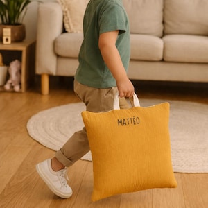 Peut inclure: Coussin carré jaune moutarde avec le nom "MATTEO" imprimé en noir. Le coussin a une surface texturée et des poignées blanches. Une personne tient le coussin en marchant.