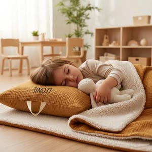 Peut inclure: Un enfant endormi repose sur un oreiller moutarde et un tapis blanc, recouvert d'une couverture assortie à l'intérieur blanc et moelleux. Le nom "LEONIE" est brodé sur l'oreiller. Un jouet en peluche blanc est tenu par l'enfant.