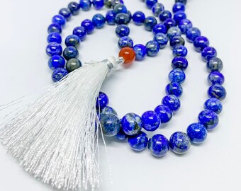 May include: Two beaded necklaces are shown. The main necklace has deep blue lapis lazuli beads, a red-orange bead, and a white tassel. A second necklace with light brown wooden beads is visible in the background.