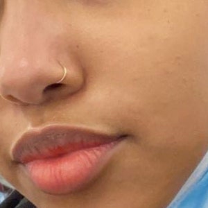 May include: Close-up of a person's face with a small gold nose ring. The person has light brown skin and is wearing a blue surgical mask.