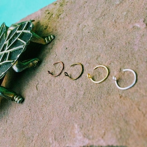 May include: Five different colored metal nose rings on a brown textured surface. The rings are arranged in a line from left to right. The rings are copper, silver, gold, and two silver rings.