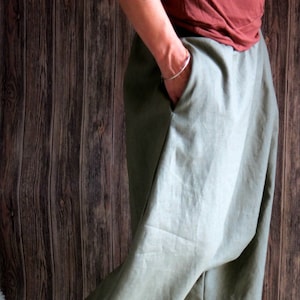 May include: Olive green linen pants with a relaxed fit. The pants have deep pockets and a wide leg. The fabric appears to be lightweight and breathable, suitable for casual wear. The pants are shown against a wooden background.