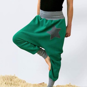 May include: A person wearing a gray beanie, a brown scarf, a black tank top, and green pants with a gray waistband and a black star. The pants are gathered at the ankles. The person is standing on a white fluffy rug. A sign with the text "ONNI PALERMO" is in the background.