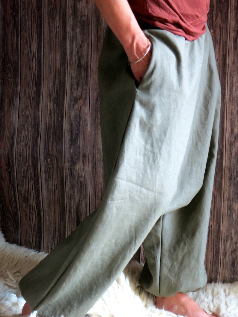 May include: Olive green linen pants with a relaxed fit. The pants have deep pockets and a wide leg. The fabric appears to be lightweight and breathable, suitable for casual wear. The pants are shown against a wooden background.