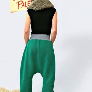 May include: A person wearing a green pair of harem pants with a gray waistband and a black shirt. The person is wearing a gray knitted hat and a brown knitted scarf. The person is standing in front of a white wall with a sign that says "ONNI PALE" in red letters.