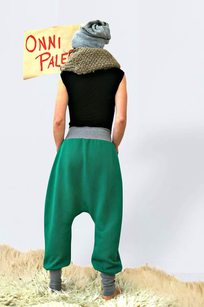 May include: A person wearing a green pair of harem pants with a gray waistband and a black shirt. The person is wearing a gray knitted hat and a brown knitted scarf. The person is standing in front of a white wall with a sign that says "ONNI PALE" in red letters.