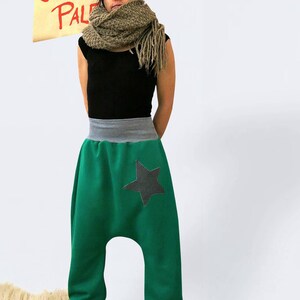 May include: A woman wearing a green pair of  harem pants with a black star on the right leg. She is also wearing a black tank top, a brown scarf, and a gray beanie. The text "ONNI PAL" is visible in the background.
