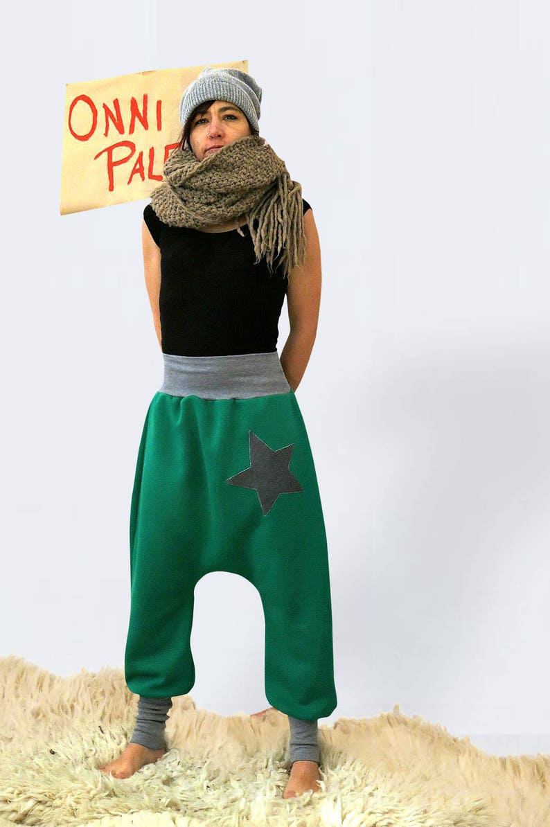 May include: A woman wearing a green pair of  harem pants with a black star on the right leg. She is also wearing a black tank top, a brown scarf, and a gray beanie. The text "ONNI PAL" is visible in the background.