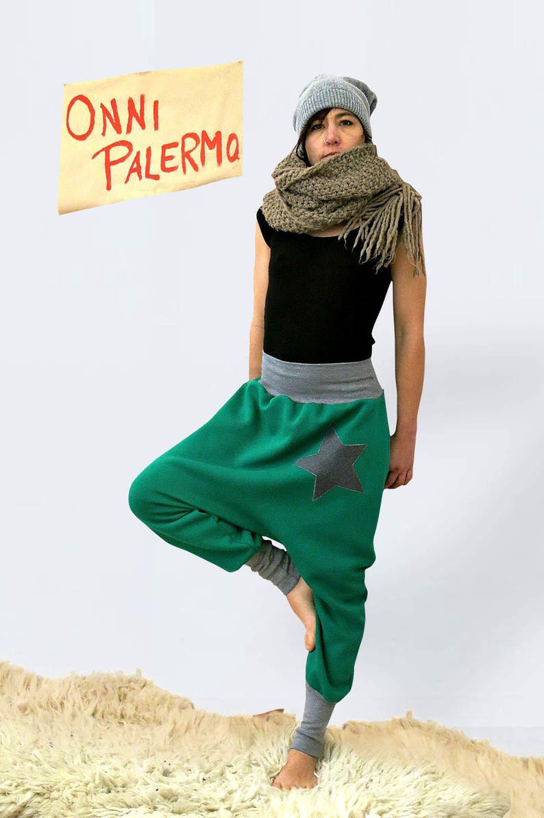 May include: A person wearing a gray beanie, a brown scarf, a black tank top, and green pants with a gray waistband and a black star. The pants are gathered at the ankles. The person is standing on a white fluffy rug. A sign with the text "ONNI PALERMO" is in the background.