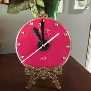 May include: A decorative clock with a vibrant pink face, adorned with small, sparkling embellishments. The clock hands are black, and the hour and minute hands are white. It's supported by an ornate, gold-coloured stand. The text "bv3" is printed on the clock face.