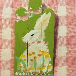 May include: A white rabbit painted on three green popsicle sticks, surrounded by yellow and orange flowers. The rabbit is sitting in a field with a pink ribbon and white dots.