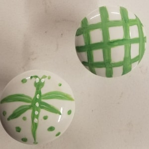 May include: Two white ceramic knobs with green painted designs. One knob features a green dragonfly with white dots, and the other has a green plaid pattern.