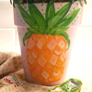 May include: A hand-painted terracotta flower pot featuring a vibrant orange and yellow pineapple design with green leaves. The pot is light pink with small green polka dots and holds a green plant. A matching pineapple fragment is in the foreground.