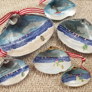 May include: Hand-painted seashell ornaments with winter scenes. Each shell features a snowy landscape with a blue sky, a Christmas tree, and a red and white striped ribbon for hanging. The ornaments are a unique holiday decoration.