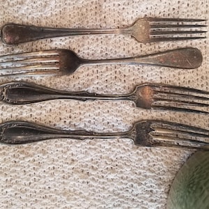 May include: Four silver forks with ornate handles. The forks are arranged in a row on a white crocheted surface.