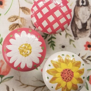 May include: Three painted wooden knobs with different designs. One knob has a red and white gingham pattern, another has a white daisy on a pink background, and the last one has a yellow sunflower on a white background.