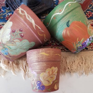May include: Three terracotta flower pots, hand-painted with botanical and autumnal designs. One pot features a pumpkin, another has leaves and berries, and the smallest pot displays leaves and grapes. The pots are arranged on a fringed textile.