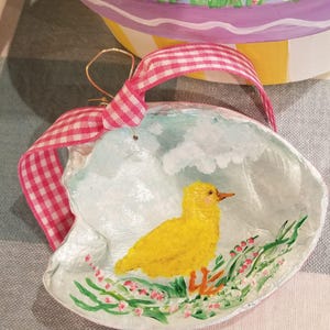 May include: A hand-painted seashell ornament with a yellow chick sitting in a bed of pink and green flowers. The ornament is tied with a pink and white gingham ribbon.
