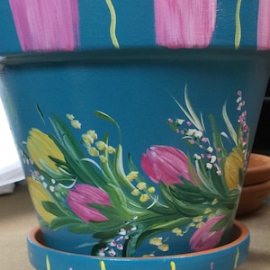 May include: A teal blue ceramic planter with a painted design of pink and yellow tulips and green leaves. The planter has a white rim and a matching saucer.