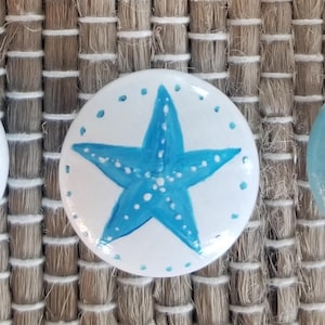 May include: Three round, white painted stones with blue designs. The first stone has blue stripes, the second has a blue starfish, and the third has a blue whale with white clouds and dots.