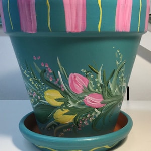 May include: A teal-colored ceramic flower pot with a painted design of pink and yellow tulips and green leaves. The pot has a white rim and a yellow and pink striped pattern around the top.