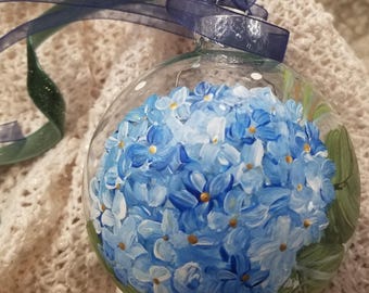Puede incluir: Una bola de Navidad de cristal transparente pintada a mano con un diseño de flor de hortensia azul. La flor tiene diferentes tonos de azul con detalles dorados. Una cinta azul oscuro está unida a la parte superior de la bola para colgarla.