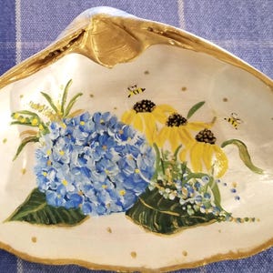 May include: A white seashell painted with a blue hydrangea, yellow flowers, and bees. The seashell has a gold rim.