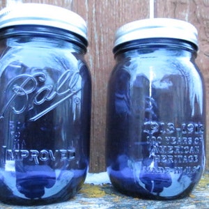 May include: Two vintage blue Ball mason jars with silver lids. The jars are embossed with the Ball logo and text. One jar reads "1913-1913 100 Years of American Heritage Made in USA". The jars are on a wooden surface.