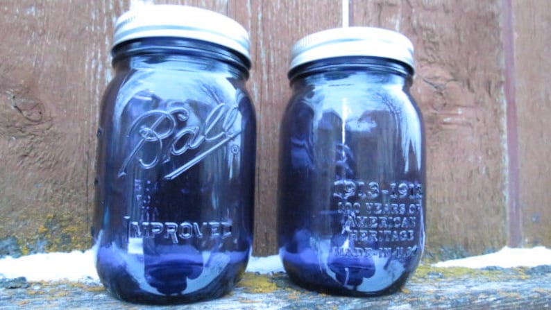 May include: Two vintage blue Ball mason jars with silver lids. The jars are embossed with the Ball logo and text. One jar reads "1913-1913 100 Years of American Heritage Made in USA". The jars are on a wooden surface.