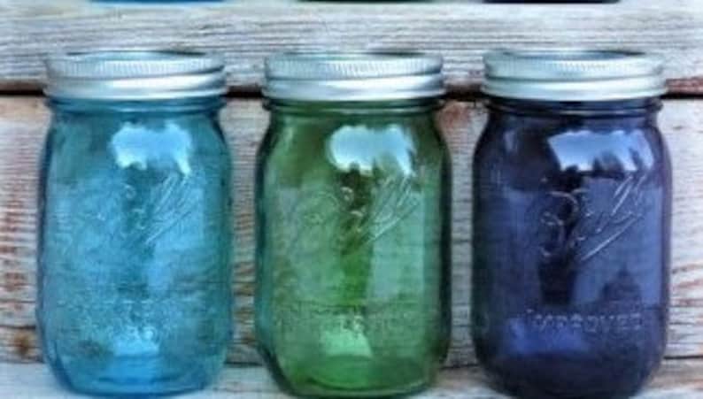 May include: Three vintage-style mason jars in shades of blue, green, and purple, each with a silver lid. The jars are arranged in a row against a weathered wooden background. The jars are approximately 6 inches tall.