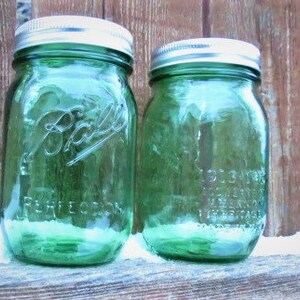 May include: Two green Ball mason jars with silver lids. The jars are embossed with the Ball logo and the word "PERFECTION". The jars are sitting on a wooden surface.