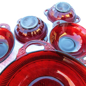 May include: A collection of ruby red glass serving dishes. The set includes several bowls with handles and a larger serving platter. The glass has a ribbed design and a glossy finish, reflecting light. The dishes are arranged on a white surface.
