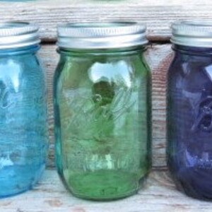 May include: Three glass mason jars in shades of blue, green, and purple. Each jar has a silver lid and embossed details. The jars are arranged side by side, showcasing their vibrant colors and classic design.