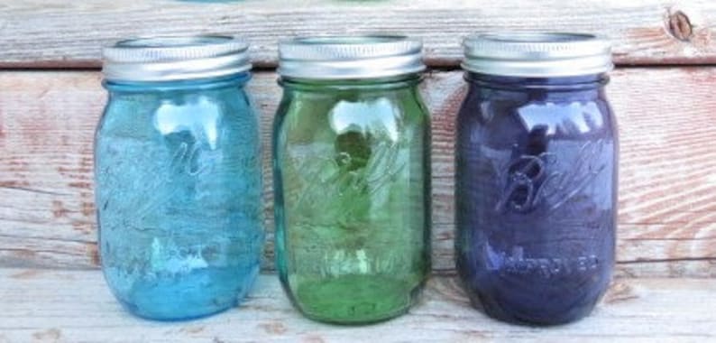 May include: Three glass mason jars in shades of blue, green, and purple. Each jar has a silver lid and embossed details. The jars are arranged side by side, showcasing their vibrant colors and classic design.