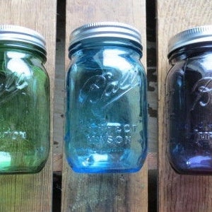 May include: Three vintage Ball mason jars in green, blue, and purple hues. Each jar has a silver lid and embossed lettering. The jars are arranged on a rustic wooden surface, showcasing their colorful glass.