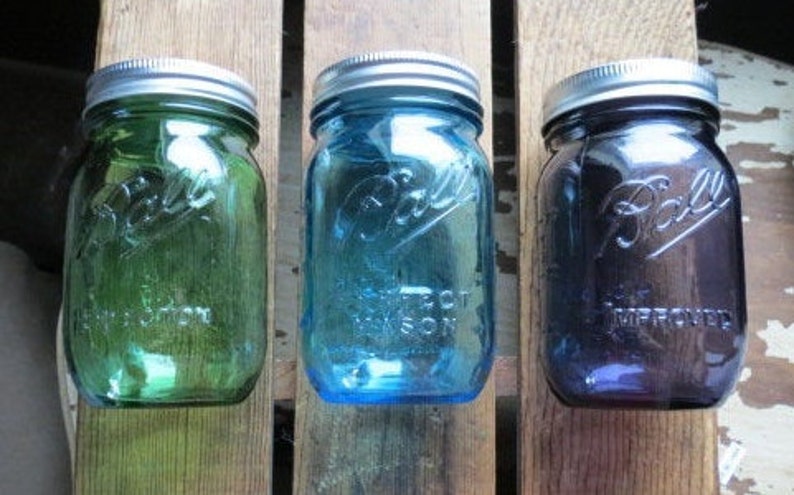 May include: Three vintage Ball mason jars in green, blue, and purple hues. Each jar has a silver lid and embossed lettering. The jars are arranged on a rustic wooden surface, showcasing their colorful glass.