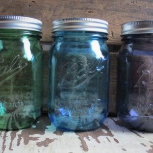May include: Three Ball mason jars in shades of green, blue, and purple. Each jar has a silver lid and the Ball logo embossed on the front. The jars are displayed on a rustic wooden surface.