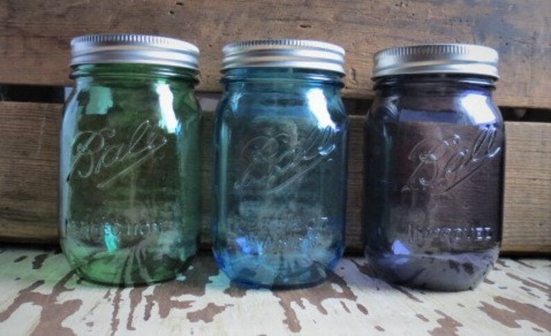 May include: Three Ball mason jars in shades of green, blue, and purple. Each jar has a silver lid and the Ball logo embossed on the front. The jars are displayed on a rustic wooden surface.