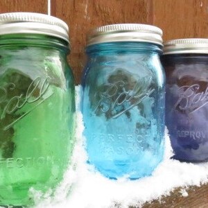 May include: Three vintage Ball mason jars in green, blue, and purple, each with a silver lid. The jars are arranged on a wooden surface covered in snow. The jars are embossed with the Ball logo and the words "PERFECT MASON".