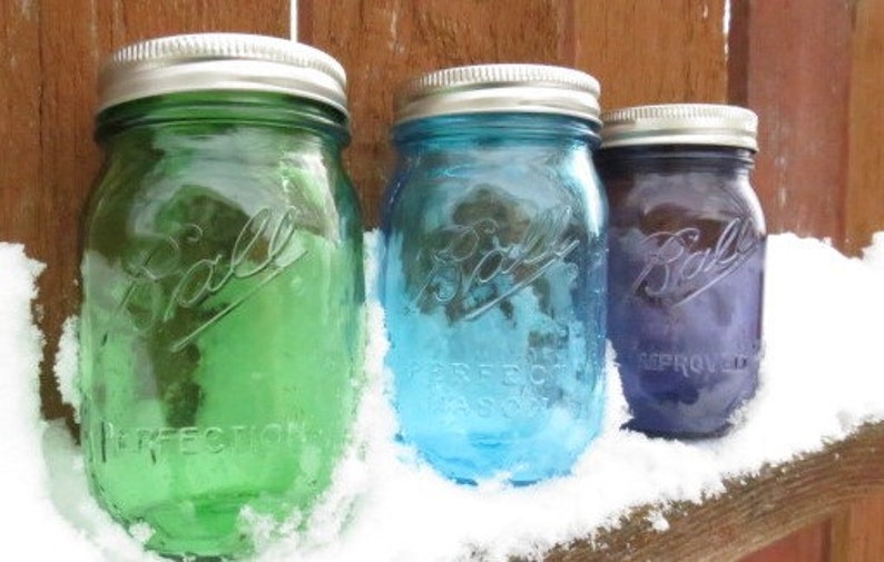 May include: Three vintage Ball mason jars in green, blue, and purple, each with a silver lid. The jars are arranged on a wooden surface covered in snow. The jars are embossed with the Ball logo and the words "PERFECT MASON".