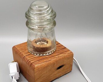 May include: A small table lamp made from a repurposed glass insulator and a wooden base. The lamp has a white cord and plug.