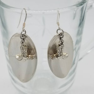 May include: Silver tone earrings with a boot charm hanging from each earring. The earrings are oval shaped and have a shiny finish.