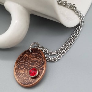 May include: A copper pendant necklace with a red gemstone and a design of an American flag. The necklace is on a silver chain.