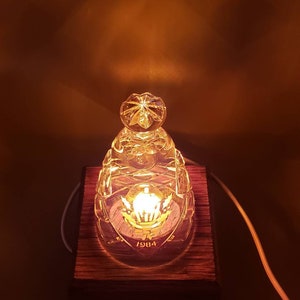 May include: A clear glass lamp base with a crown design and the year 1984 etched on it. The lamp base is sitting on a wooden base.